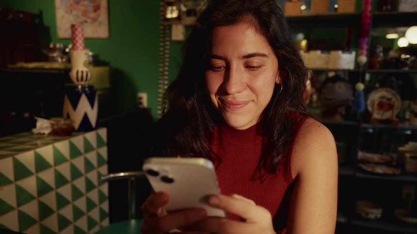 A smiling young woman texts on her smartphone while sitting in a cozy, eclectic cafe with warm lighting