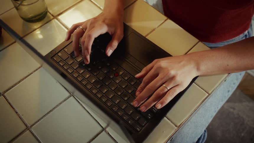 An overhead shot of a woman's hands typing on a laptop keyboard placed on a tiled kitchen counter