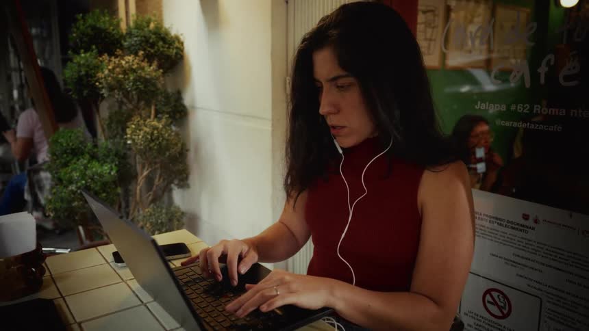 A focused young woman wearing earbuds works diligently on her laptop at an outdoor cafe table