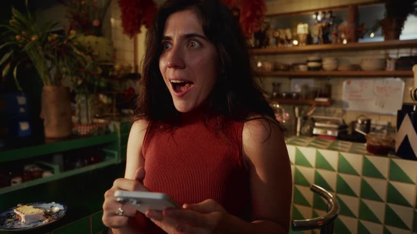 A woman's focused expression turns to one of pure shock and excitement as she looks at her phone in an eclectic cafe