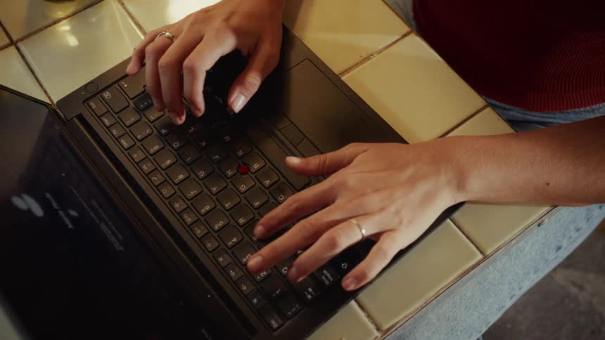 Overhead close-up of a person's hands typing on a laptop keyboard on a tiled cafe table