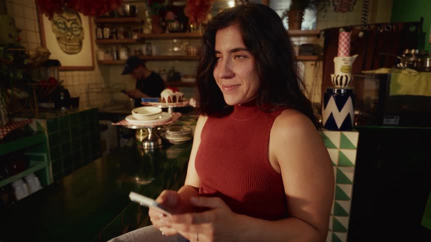 A woman smiles while using her smartphone, sitting at the counter of a quirky and colorful cafe in the evening