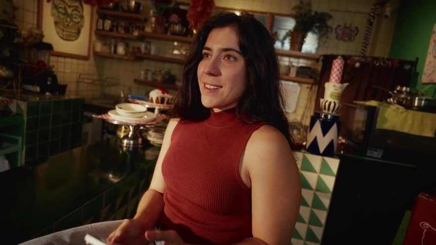 A young woman scrolls on her smartphone before looking up with a warm smile in a quirky, art-filled kitchen
