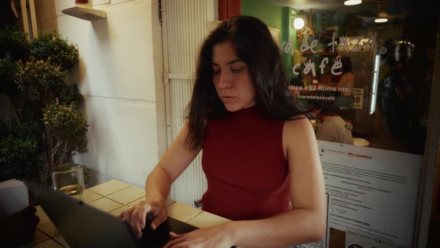 A young woman opens her laptop and begins working at a small table outside a bustling city cafe at night