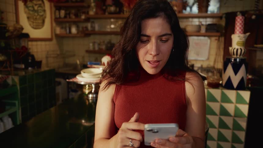 A woman sitting at a counter in a funky kitchen has a look of surprised delight while scrolling on her smartphone