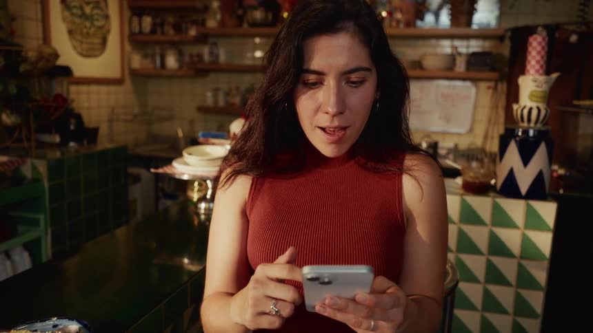 A young woman in a red top scrolls through her smartphone while sitting at the counter of a quirky, warmly lit cafe