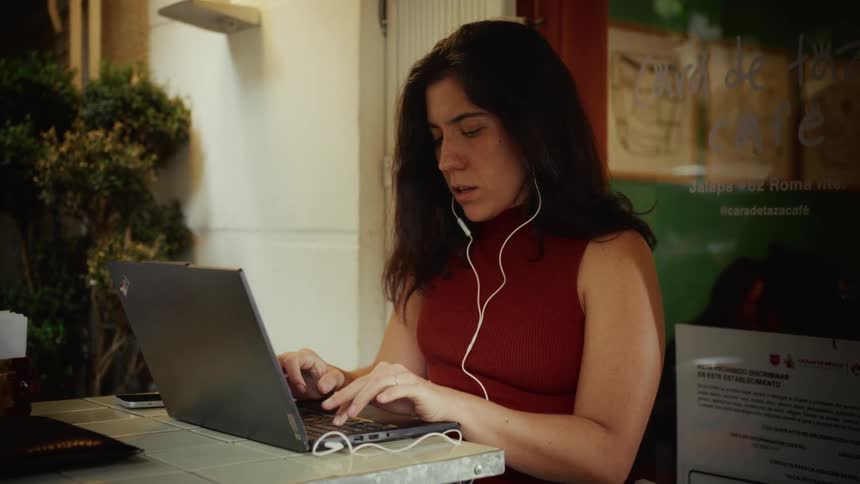 Focused woman wearing earphones works on her laptop at an outdoor table of a cafe in the evening