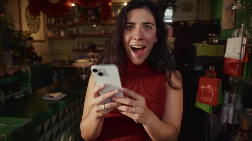 A woman looks up from her smartphone with a shocked and happy expression while standing in an eclectic shop