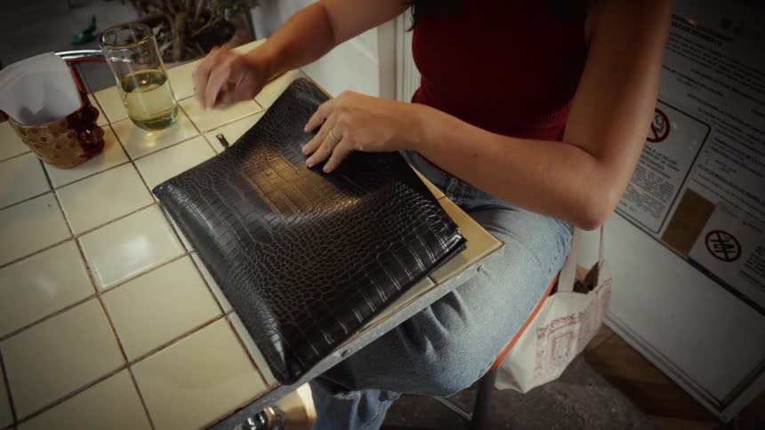 A woman sits at a cafe table and slowly unzips a black alligator-textured laptop case