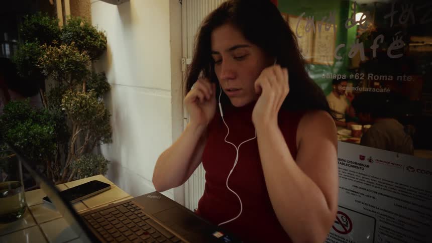 A woman sitting outside a cafe at night puts in her earphones before starting to work on her laptop