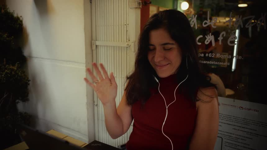 A smiling woman wearing earbuds waves to her laptop screen during a video call at an outdoor cafe