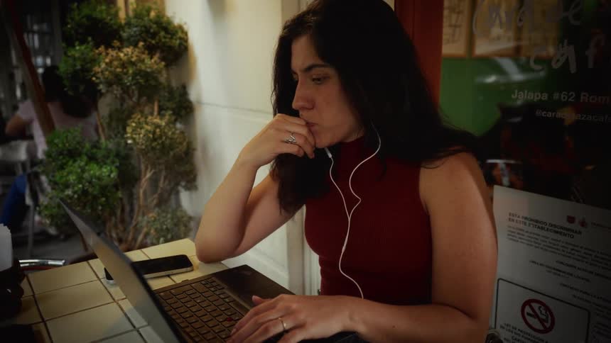 A focused woman with earbuds works on her laptop in slow motion at an outdoor cafe table
