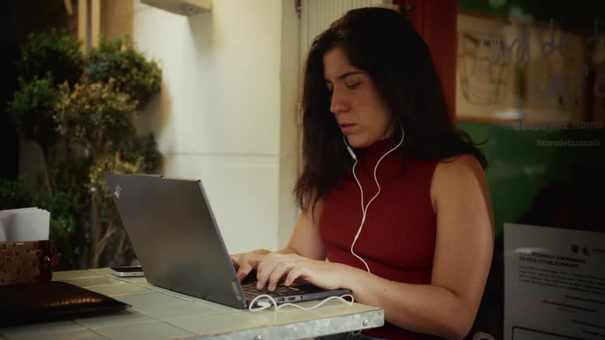 A focused woman with earbuds works diligently on her laptop at an outdoor cafe table at night