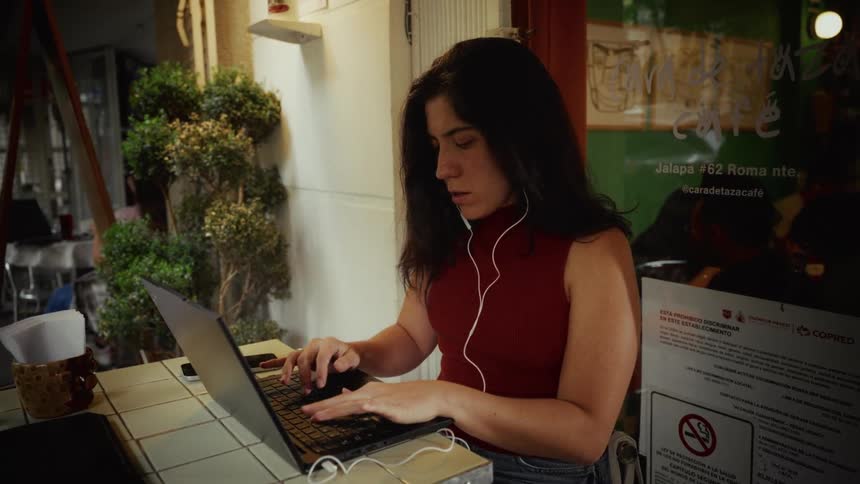 A focused woman with earphones types on her laptop while sitting at an outdoor cafe table