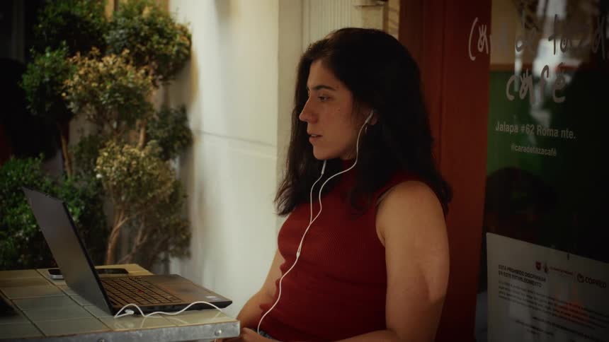 A focused woman with earbuds works on her laptop at an outdoor city cafe, then pauses to watch the screen