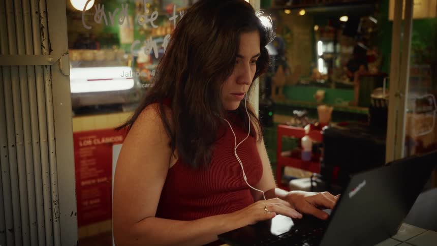 A focused woman with earbuds diligently types on her laptop while working remotely from a colorful cafe