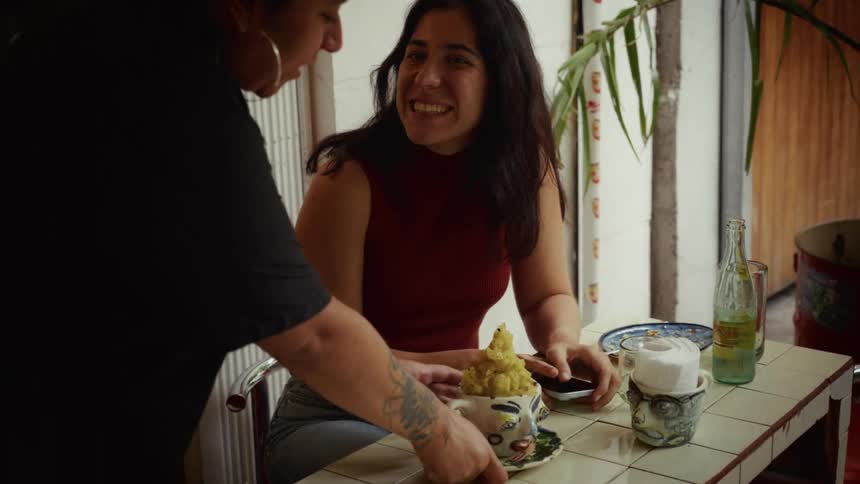 A woman at a cafe table is pleasantly surprised when a waitress serves her a unique drink in an artistic, face-shaped mug, causing her to smile and laugh