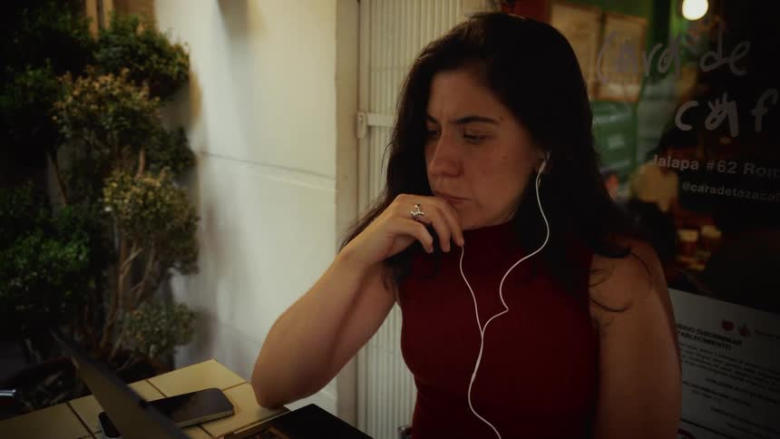 A woman with headphones intently works on her laptop at an outdoor cafe table at night, pausing to think