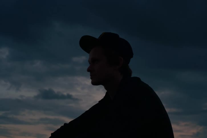 Silhouette of a young man in a backward cap looking pensive against a dark and cloudy sky at twilight