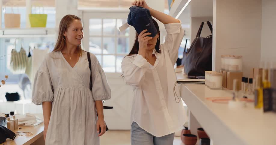 Two female friends have a fun day shopping together at a stylish clothing boutique, trying on a baseball cap
