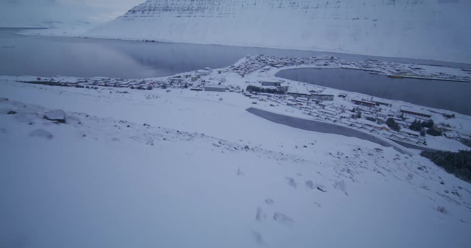 A dynamic first-person perspective shot moves over a snowy mountain to reveal a picturesque fishing village in a tranquil fjord