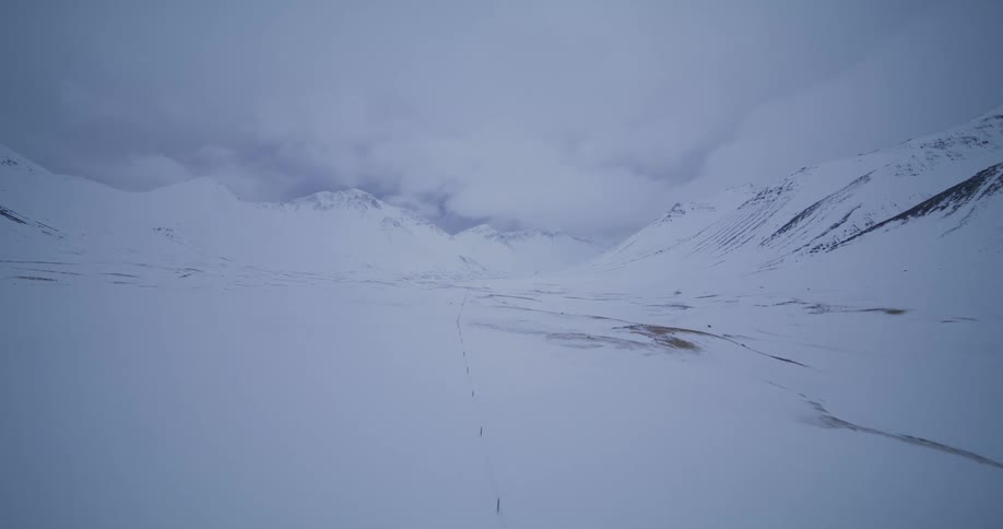 Aerial drone footage flies over a vast, desolate snow-covered valley between mountains on a bleak winter day