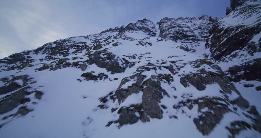 An FPV drone flies up the steep, rocky face of a vast, snow-covered mountain