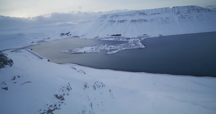 An epic aerial reveal flying out from a steep, snowy mountain pass over a remote coastal fjord town in winter