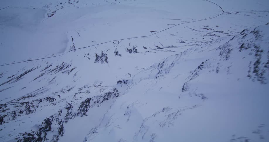 A dramatic first-person view drone speeds through a rugged canyon in a vast, snow-blanketed mountain landscape