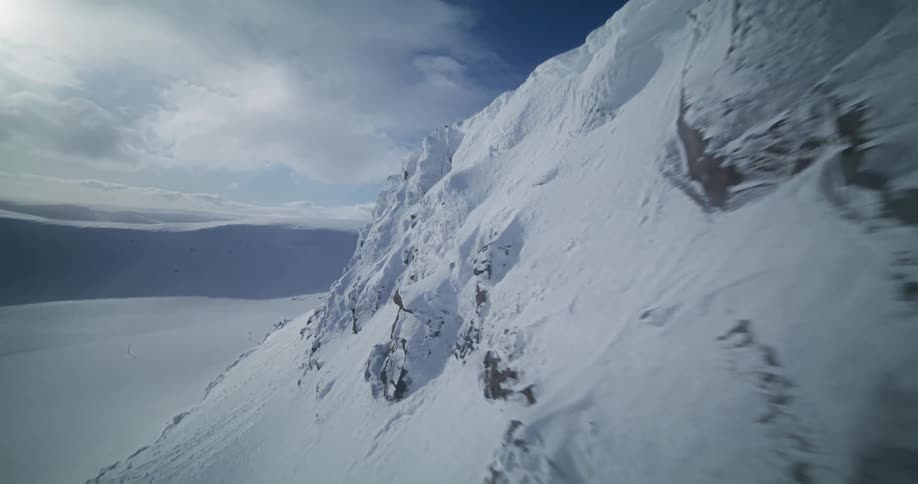 A fast-paced aerial shot glides along the precipice of a snow-covered mountain on a bright winter day