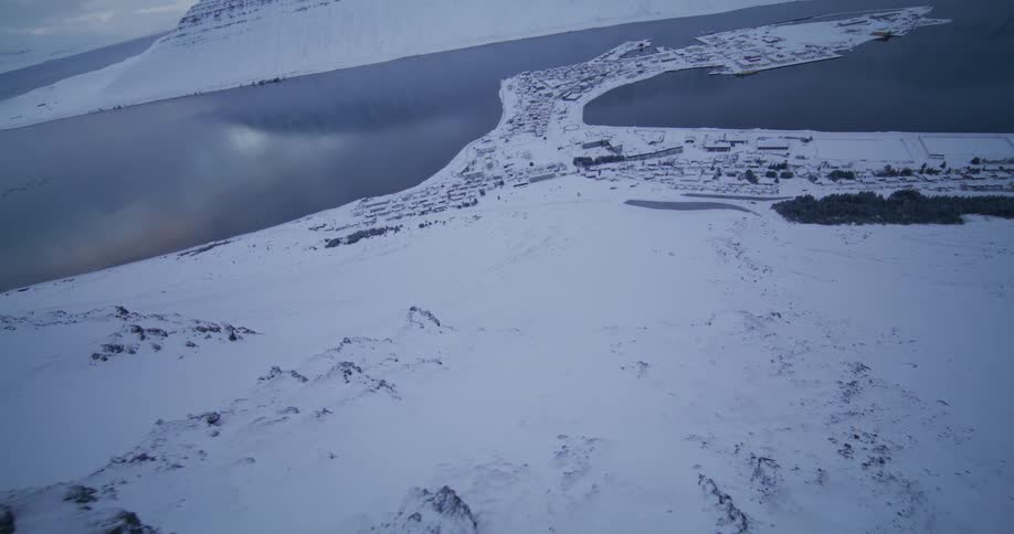 A stunning aerial shot flies over a snowy mountain ridge revealing a remote coastal town nestled in a vast fjord during winter