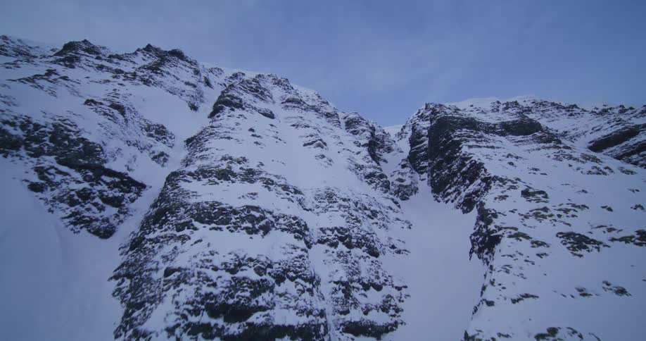 A dramatic aerial ascent up a formidable, snow-covered mountain towards a narrow pass under a winter sky