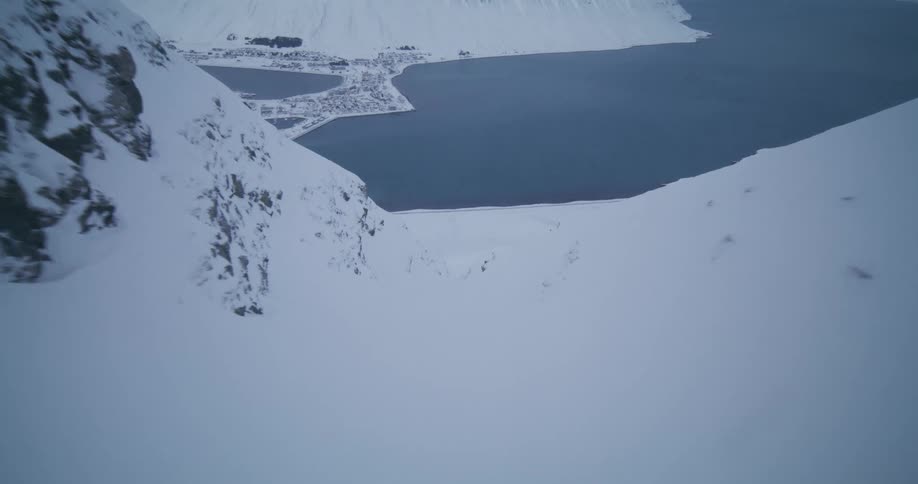 First-person perspective flying down a steep, snowy mountain couloir towards a coastal town nestled in a fjord