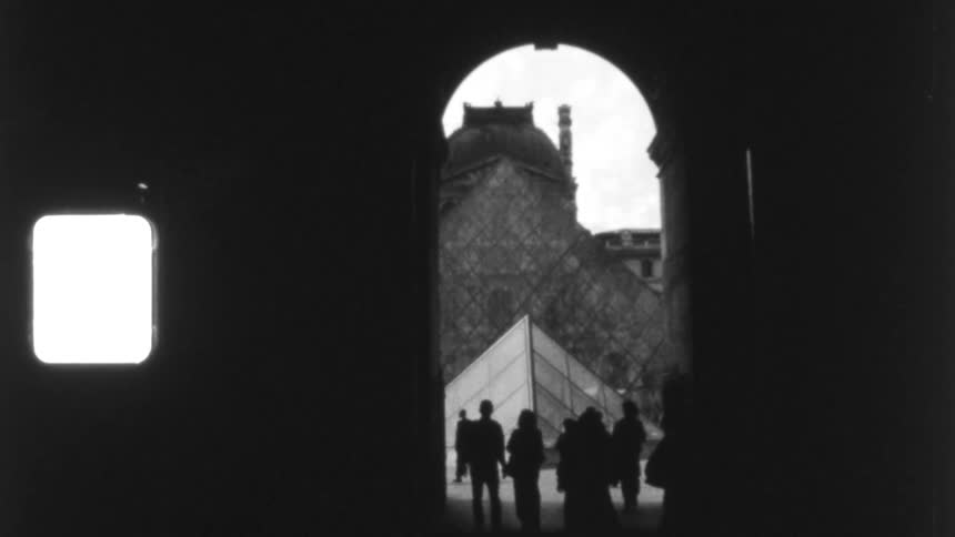 A vintage, black and white shot shows silhouetted people walking through an archway framing the Louvre Pyramid in Paris