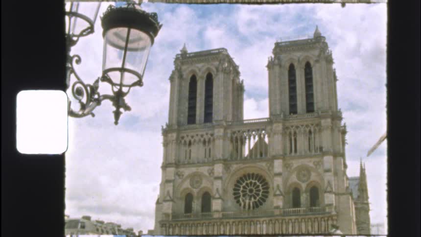 Vintage 8mm film captures a static, low-angle shot of the iconic Notre Dame Cathedral in Paris, France, with a classic street lamp in the foreground