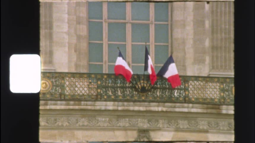 Vintage film captures the French Tricolore flags waving gently from an elegant, classic Parisian balcony