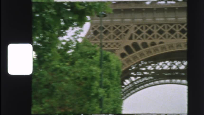 Vintage 8mm film footage captures a handheld, low-angle tour of the Eiffel Tower in Paris on an overcast day