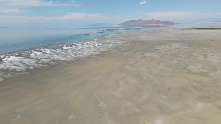 A slow aerial push-in shot reveals the vast, textured salt flats and serene waters of a salt lake shoreline with distant mountains