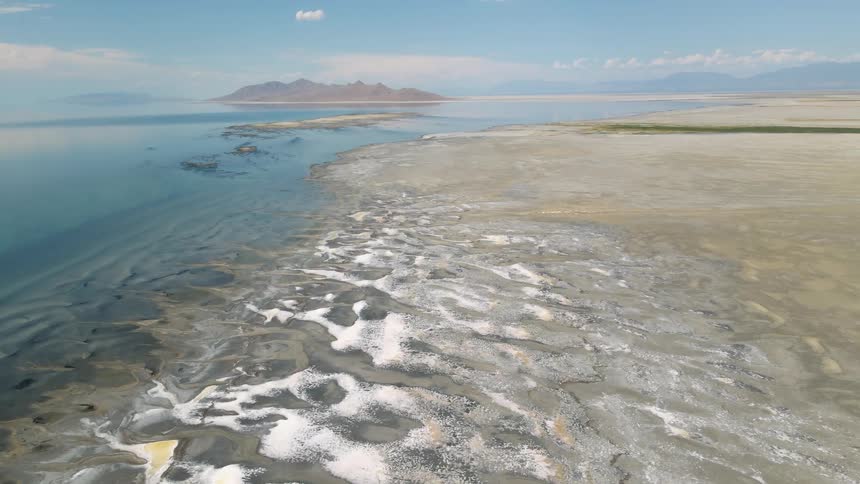 A stunning wide aerial shot captures the vast and desolate beauty of the Great Salt Lake shoreline with its calm blue water and white salt flats
