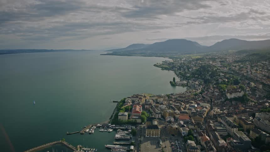A wide aerial shot pulls back to reveal a beautiful European city and marina on the coast of a lake, with mountains under a cloudy sky