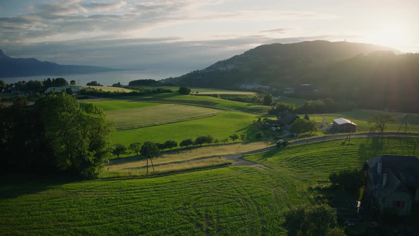 A tractor drives through the idyllic green countryside with a lake and mountains in the background during a beautiful sunset