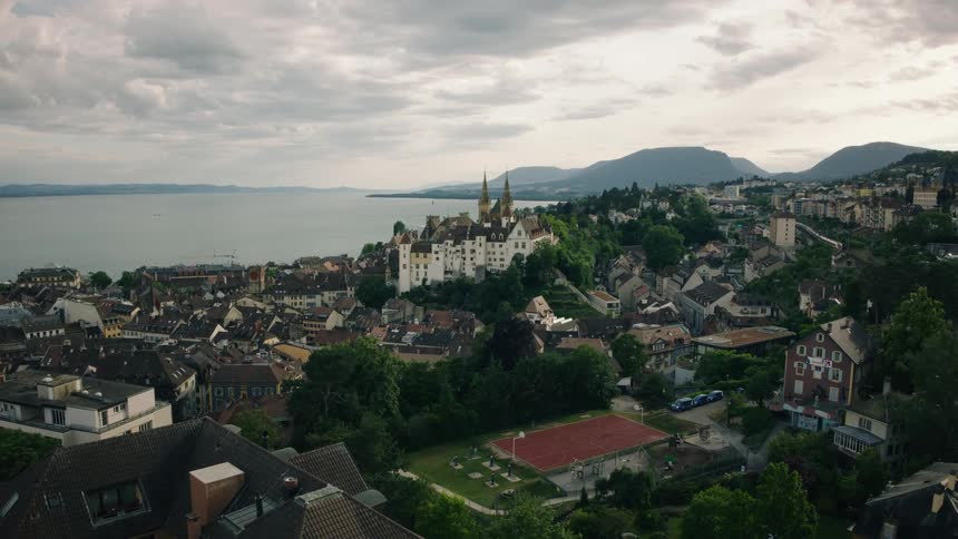 A picturesque European town and its historic castle sit on the shores of a vast lake under a dramatic cloudy sky