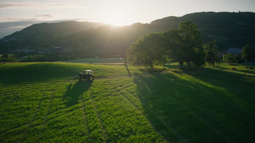 Aerial view of a tractor driving through a green field on a picturesque farm at sunset