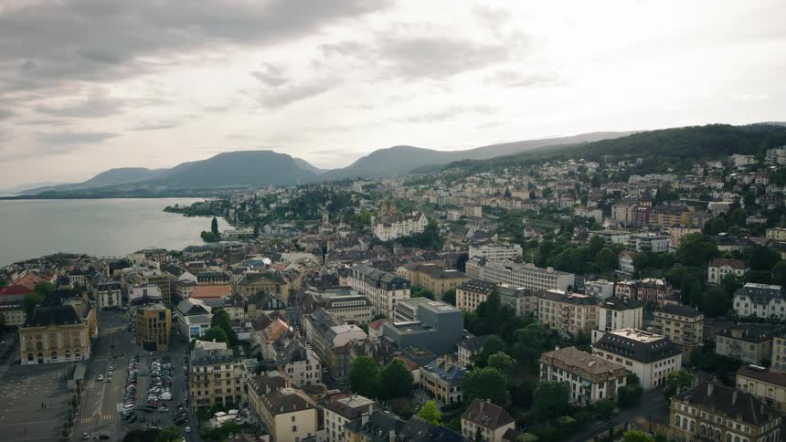 A static wide aerial view of the historic city of Neuchâtel, Switzerland, nestled on the shores of a vast lake with rolling green hills in the background