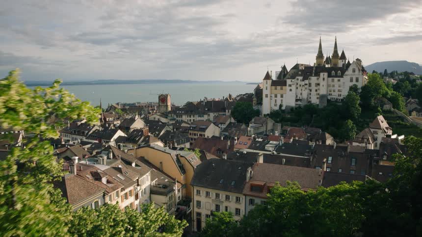 An aerial shot slowly pushes over green trees to reveal a picturesque historic European town and castle on the shore of a vast lake