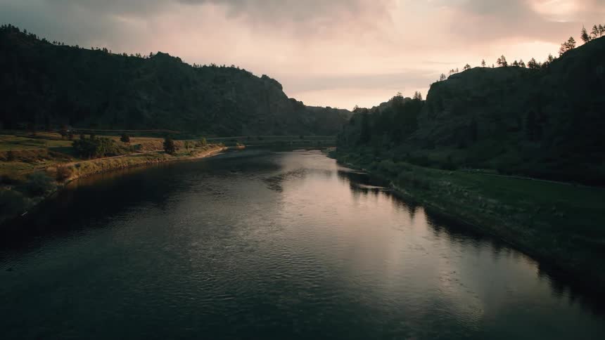 An atmospheric drone shot slowly flies over a steel bridge and down a tranquil river canyon at dusk