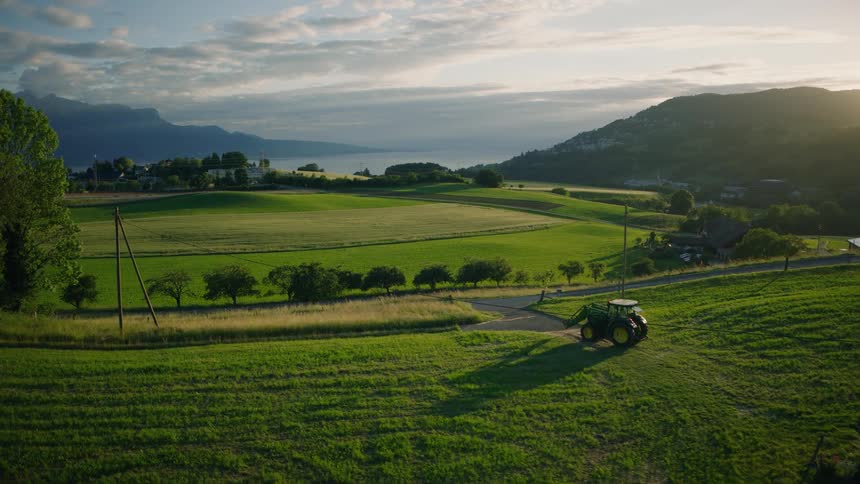 An aerial view captures a tractor driving through a vibrant green countryside landscape at sunset with a lake and mountains in the background