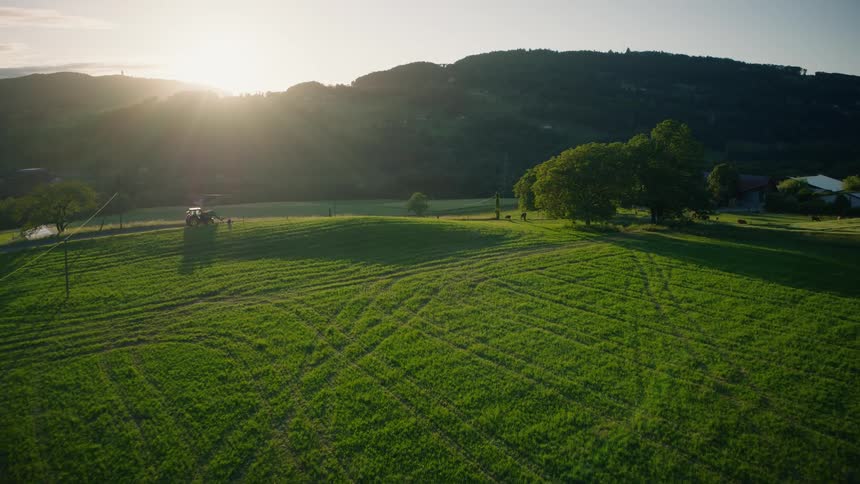 An aerial shot captures a tractor working on a farm at sunset amidst rolling green hills
