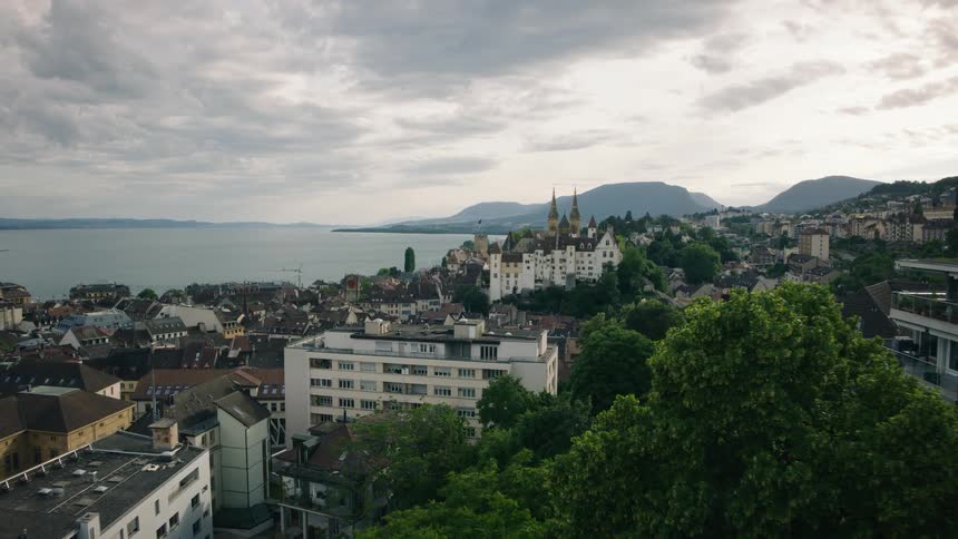 A high-angle pull-back shot reveals the historic city of Neuchâtel, Switzerland, with its castle overlooking the lake on an overcast day