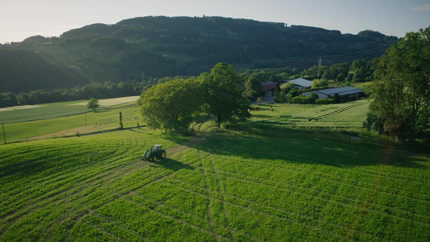 Idyllic aerial view of a tractor cultivating a lush green field on a farm nestled in rolling hills at sunset
